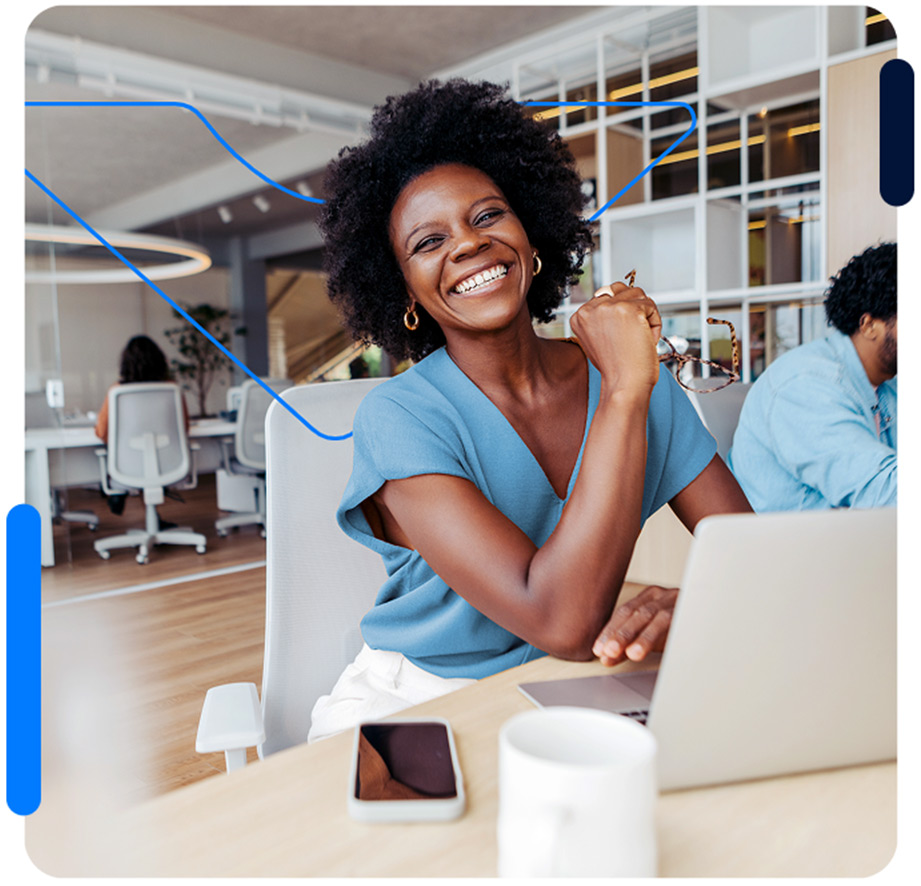Mulher sentada, sorrindo, em frente de notebook e celular, dentro de escritório