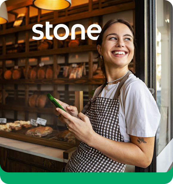 Mulher em pé, sorrindo, trabalhando em padaria e segurando celular. Ao fundo, está escrito 'Stone'